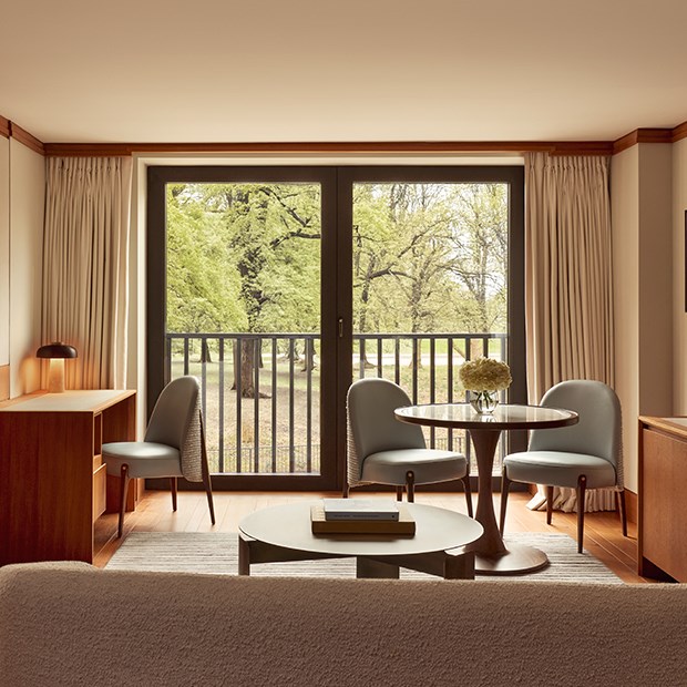 Bright suite interior with a small dining table, pale blue chairs, and a writing desk, overlooking a park through large glass doors.