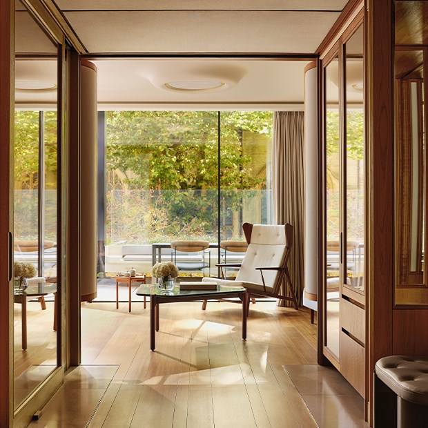 Warm, wood-accented living space with a modern lounge chair and glass coffee table, opening onto a sunlit terrace framed by trees in early autumn colours.