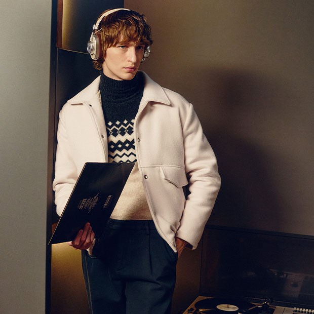A young man is wearing a light jacket over a patterned turtleneck sweater and listening to music with large over-ear headphones while holding a vinyl record. He is standing in an indoor setting, likely a music studio, with a record player visible in the background.
