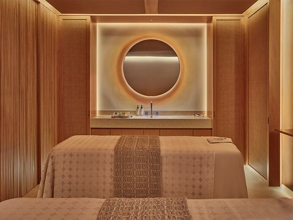 A serene spa treatment room with two massage tables, soft lighting, and a circular mirror above a minimalist vanity.