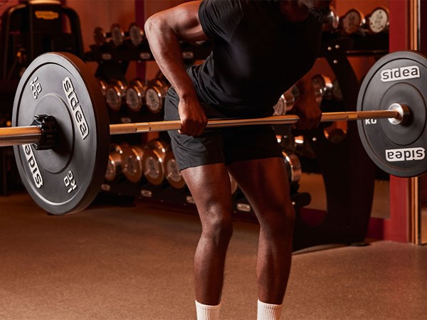 A man in athletic clothing performs a barbell row in a gym, with weight racks visible in the background.