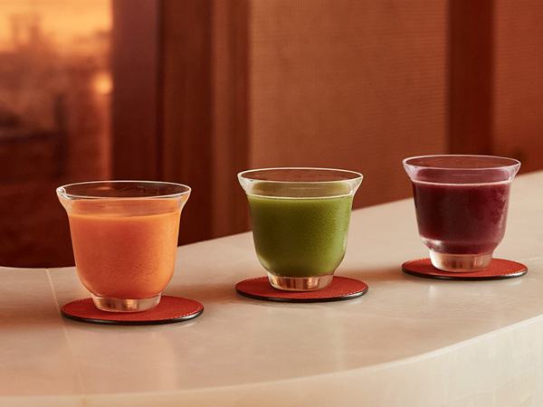 Three colourful wellness drinks—orange, green, and deep red—served in glass cups on coasters atop a curved stone counter.