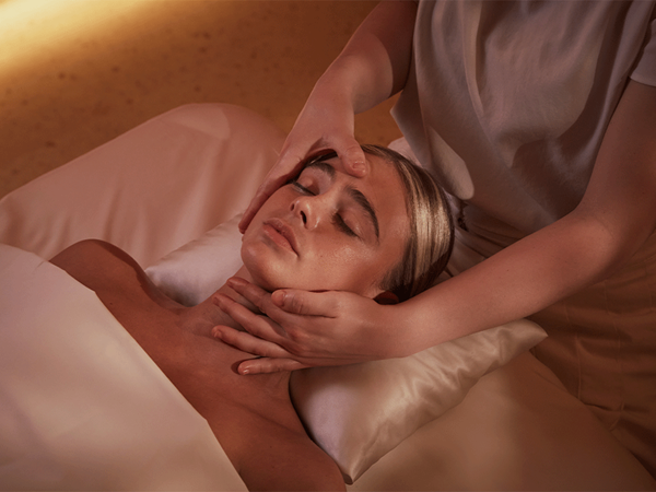 A woman receives a relaxing facial massage while lying on a treatment bed, with warm lighting creating a calming atmosphere.