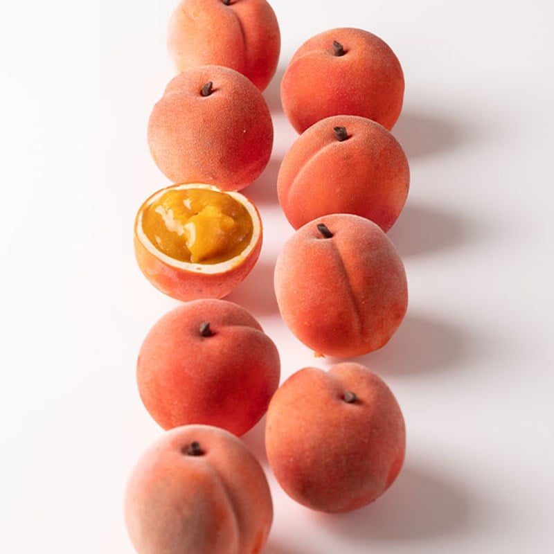 Realistic peach-shaped desserts with velvety skin arranged in a row, one halved to reveal juicy golden fruit filling inside.