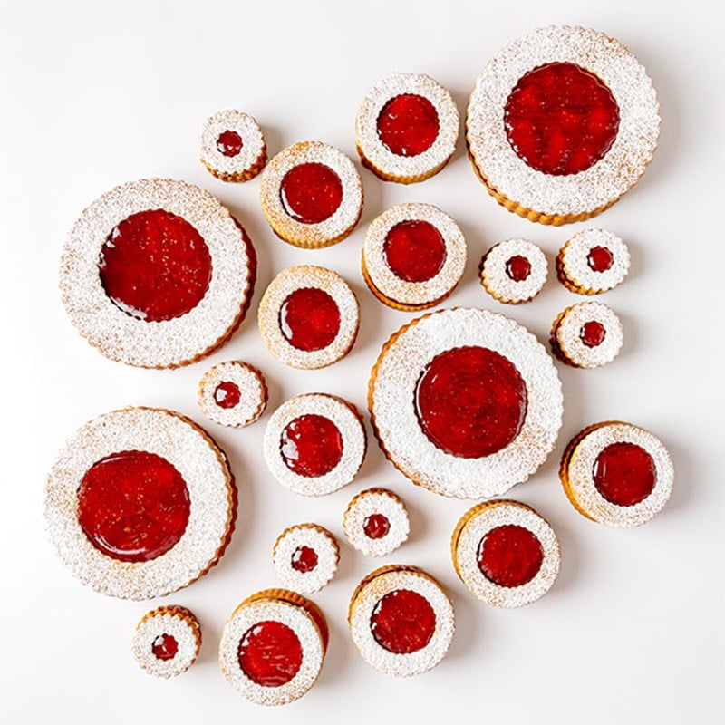 A collection of Linzer-style cookies with red jam centres and powdered sugar rings, displayed in various sizes against a white background.