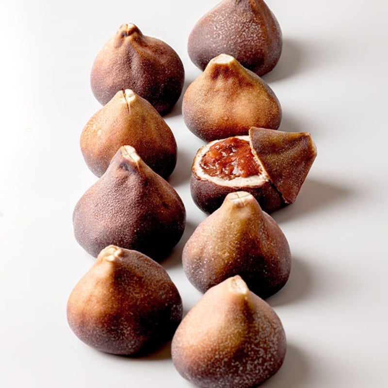 Lifelike fig-shaped pastries arranged in rows, with one cut open to show a rich, dark fruit filling inside a realistic skin.