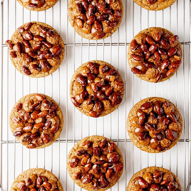 Twelve round praline cookies topped with glossy caramelised nuts, evenly spaced on a wire cooling rack against a bright white surface.