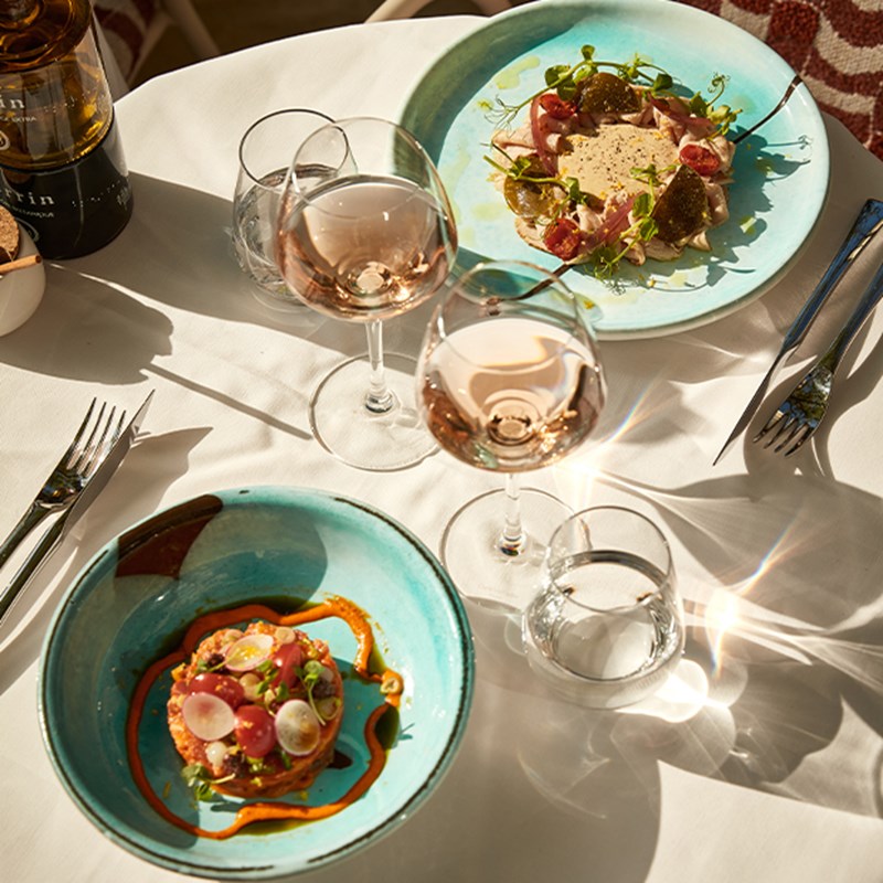 Overhead view of two colorful gourmet plates with rosé wine glasses on a sunlit white tablecloth set for fine dining.