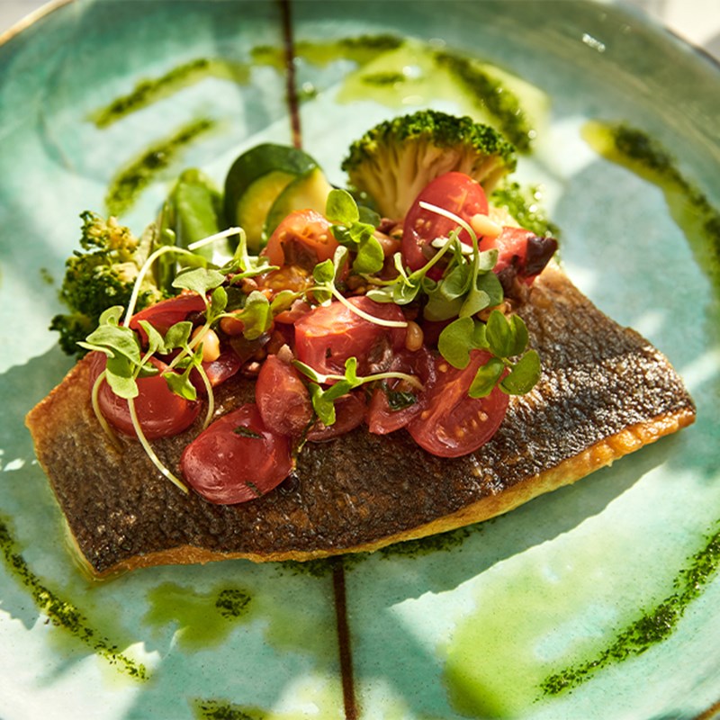 Plated dish featuring seared fish fillet topped with cherry tomatoes and microgreens, served with broccoli and zucchini on a green ceramic plate.