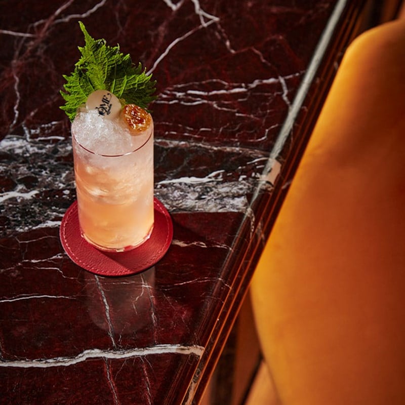 A refined cocktail served over crushed ice, garnished with fresh shiso leaves, caviar, and a bespoke branded sphere, placed on a rich marble countertop.
