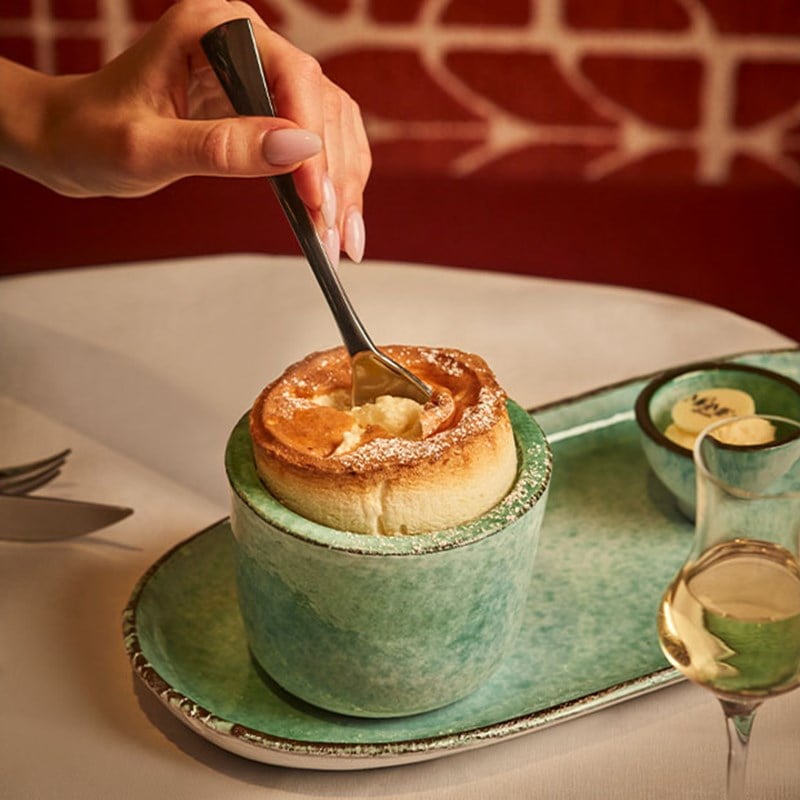 A delicate hand elegantly breaks into a golden soufflé, its airy texture revealed beneath a dusting of powdered sugar, served in a rustic green ceramic dish alongside a glass of fine wine.