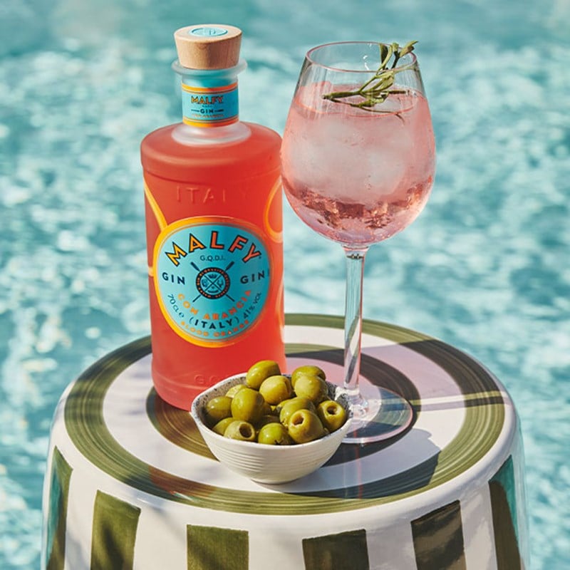 Malfy gin bottle, pink cocktail with rosemary, and green olives by a poolside.