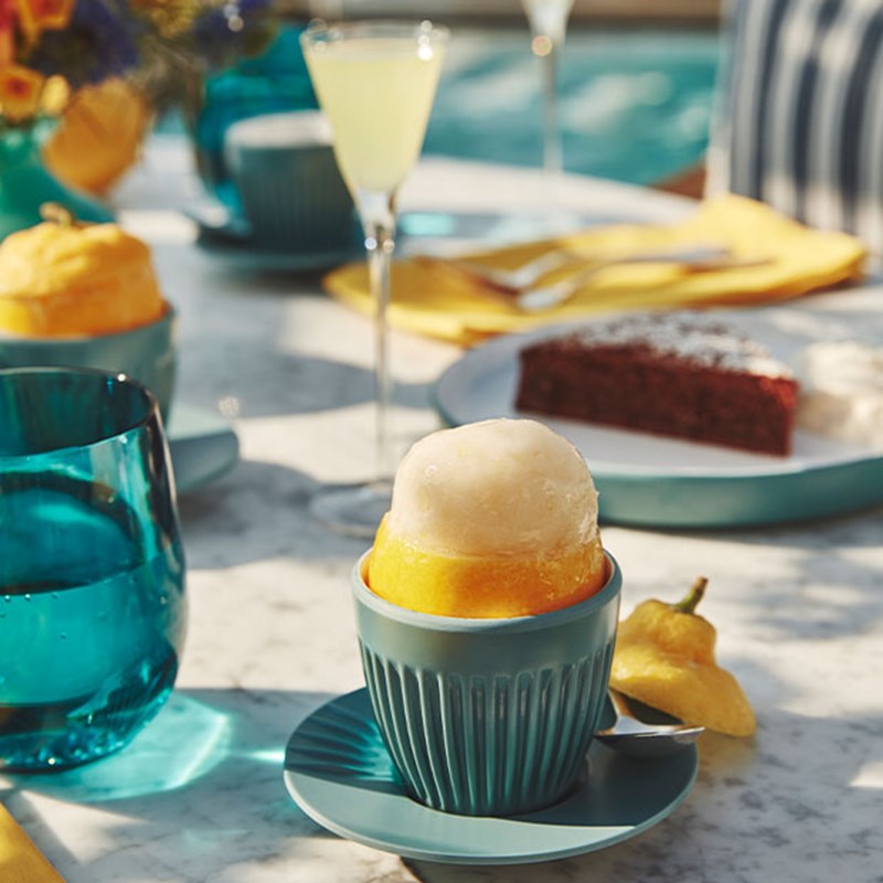 Lemon sorbet served in a hollowed lemon on a blue cup, with desserts and limoncello in the background.