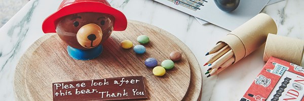 Paddington Bear cake with smarties and note made from chocolate, colouring pencils, London book, games console controller and children's activity book on table.