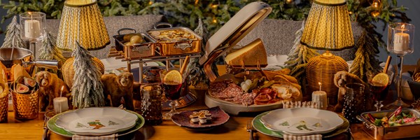 A lavish holiday dining setup featuring copper cookware, a variety of cured meats, cheeses, bread, and festive decor, complete with candlelight, miniature snowy trees, and warm lamps, creating a cozy Christmas ambiance.