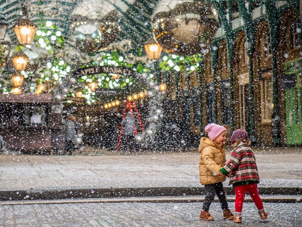 Two young children in warm winter coats and hats joyfully play together in the falling snow under the festive lights and decorations of Covent Garden’s Apple Market.