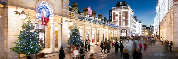 A festive evening scene at Covent Garden in London, with illuminated buildings, Christmas trees adorned with lights, and people strolling along the cobblestone streets.