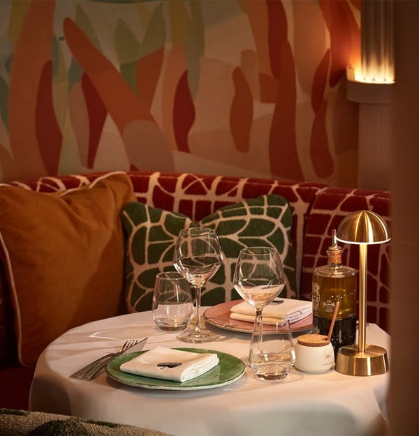 A warmly lit dining table set against a backdrop of abstract wall art, featuring plush seating, elegant tableware, and ambient golden lighting.