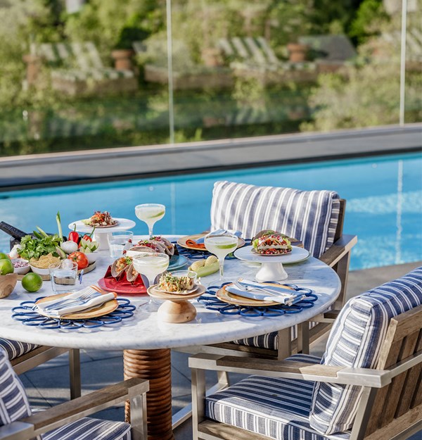 Round table with a spread of mexican food including tacos and margaritas, blue striped chairs tucked underneath and view of the rooftop pool in the background.