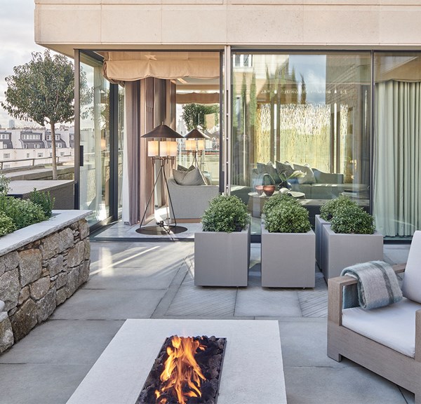 A modern rooftop terrace features a cozy seating area with a fire pit, lush greenery, and large glass doors leading to an elegant indoor living space.