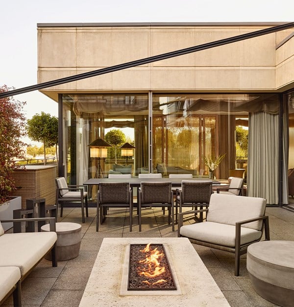 A stylish private terrace with modern cushioned seating, a rectangular stone firepit, and a dining area, framed by glass doors leading to a softly lit interior lounge.