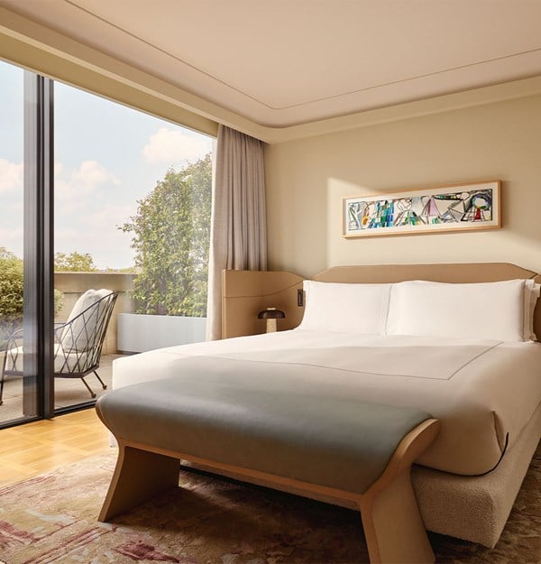 A serene bedroom with a minimalist beige bed, modern artwork, and sliding glass doors opening to a lush private terrace with outdoor seating and views of greenery.