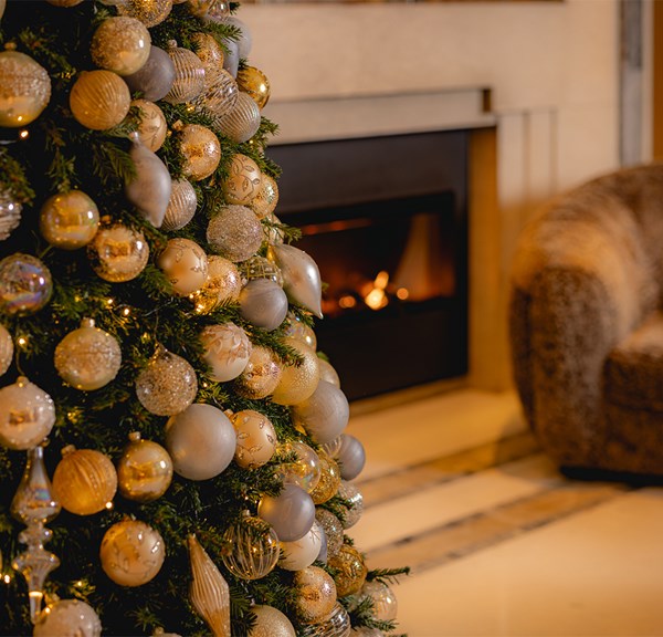 A beautifully adorned Christmas tree with gold and silver ornaments stands beside a cozy lounge chair and a lit fireplace, creating a warm and festive ambiance.