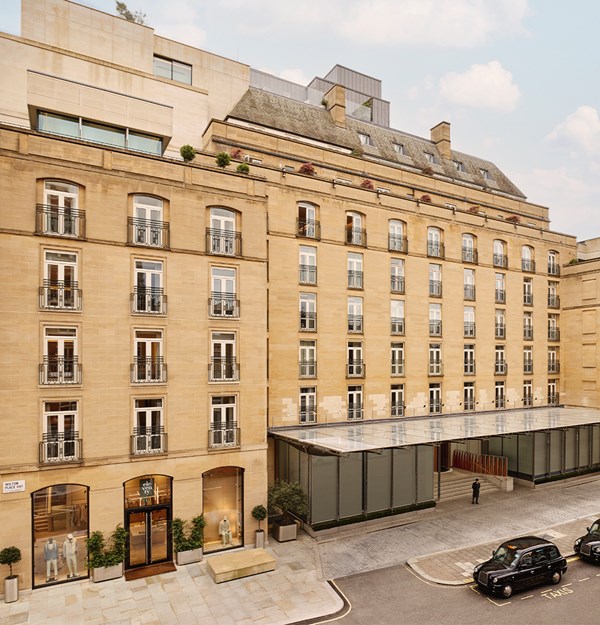 Exterior view of The Berkeley hotel in London, showcasing its elegant sandstone façade, French balconies, street-level boutique entrances, and a sleek glass canopy at the main entrance with black cabs parked outside.