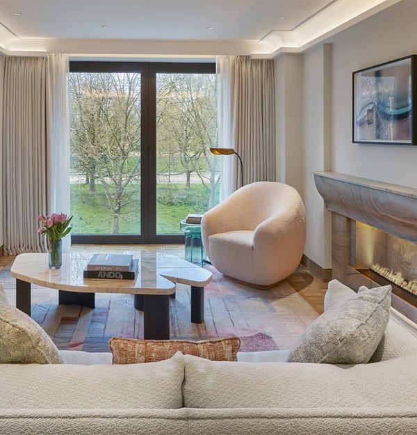 A luxurious modern suite with a cozy living area, featuring a light-colored sofa, a unique marble coffee table, and a soft armchair near large windows that offer a view of greenery. The room is complemented by a sleek fireplace and an adjacent bedroom.