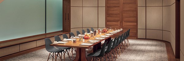 A long conference table set for a meeting, featuring black chairs and neatly arranged notepads, cups, and various snacks in jars and bowls. The room has wood-paneled walls, a frosted glass window, and a soft, patterned carpet.