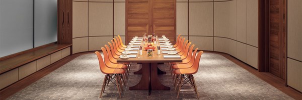 A long conference table set for a meeting, featuring orange chairs and neatly arranged notepads, cups, and various snacks in jars and bowls. The room has wood-paneled walls, a frosted glass window, and a soft, patterned carpet.