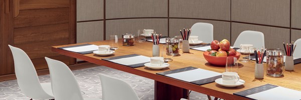 A small section of a  long conference table set for a meeting, featuring white chairs and neatly arranged notepads, cups, and various snacks in jars and bowls. The room has wood-paneled walls, a frosted glass window, and a soft, patterned carpet.