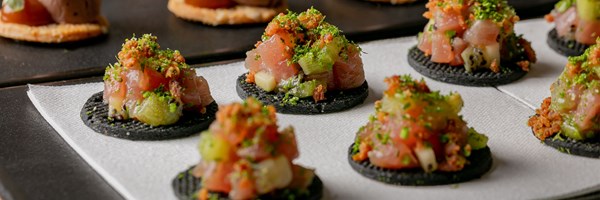 a close-up of gourmet appetizers arranged on a tray, featuring small, black crackers topped with a colorful mix of diced raw fish, green herbs, and other finely chopped ingredients. The appetizers are garnished with a sprinkle of green herbs and are served on a white napkin.