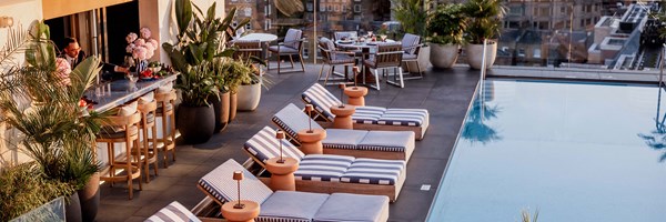 A rooftop pool area features striped lounge chairs, potted plants, and a bar with stools. In the background, there are dining tables set with meals, creating a relaxing and inviting atmosphere.