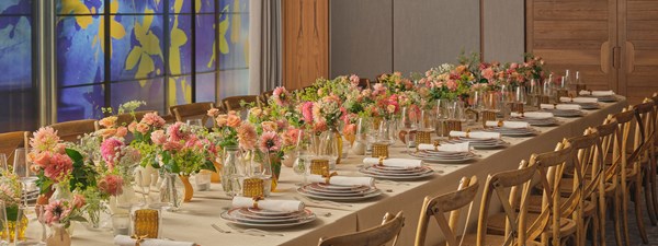 A long banquet table adorned with vibrant, colorful floral arrangements and neatly set tableware in an upscale event setting with wooden chairs.