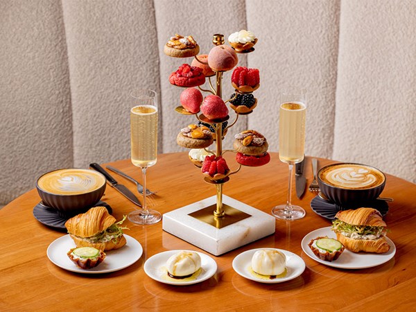 A beautifully arranged afternoon tea spread features a tiered stand with assorted pastries and sweets. The table also includes two lattes, champagne flutes, filled croissants, and creamy desserts, creating an elegant setting.