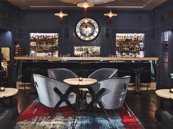 An elegant and moody bar setting featuring plush grey armchairs, a polished marble bar, illuminated shelves of premium spirits, and sophisticated décor with deep blue walls and warm ambient lighting.