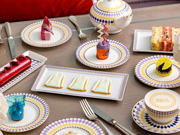 A vibrant spread of fashion-inspired pastries and sandwiches displayed on geometric-patterned china, including delicate suit-shaped biscuits and colorful desserts, paired with a teapot and fine teacups