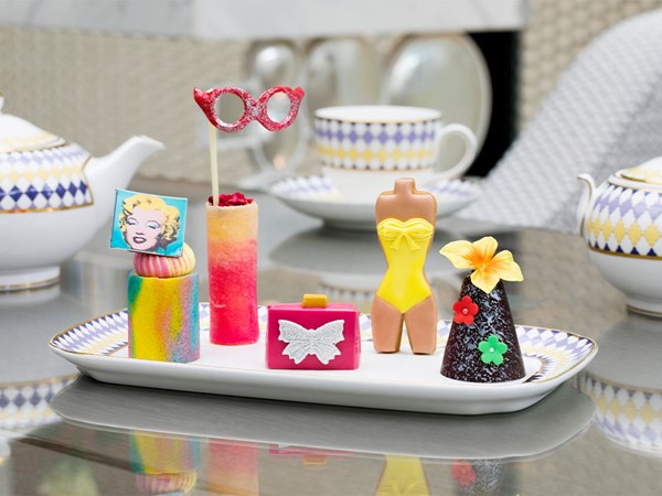 A playful and stylish dessert presentation featuring a Marilyn Monroe-themed pastry, colorful cakes, a yellow swimsuit-shaped treat, and a floral cone dessert, served on elegant patterned china.