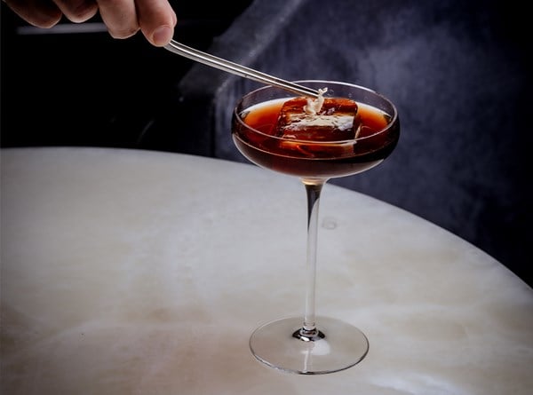 A close-up of a dark cocktail in a tall-stemmed glass with a large ice cube, as a hand carefully places a garnish on top using tweezers.