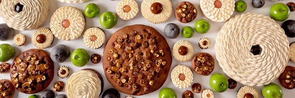 An assortment of intricately decorated pastries featuring rich textures, surrounded by green apples and a few dark fruits, arranged on a white background.