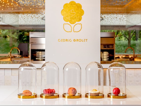 Cedric Grolet's 5 signatures for Summer 2024 lined up in a row in glass cake domes; almond, raspberry tart, peach, lemon flower, vegan strawberry.