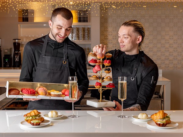 Two men in black shirts and aprons, man on the left is holding a box of patisserie, and thee man on the right is holding a tiered stand of cakes. In the image croissant sandwiches and 2 glass of champagne are featured.
