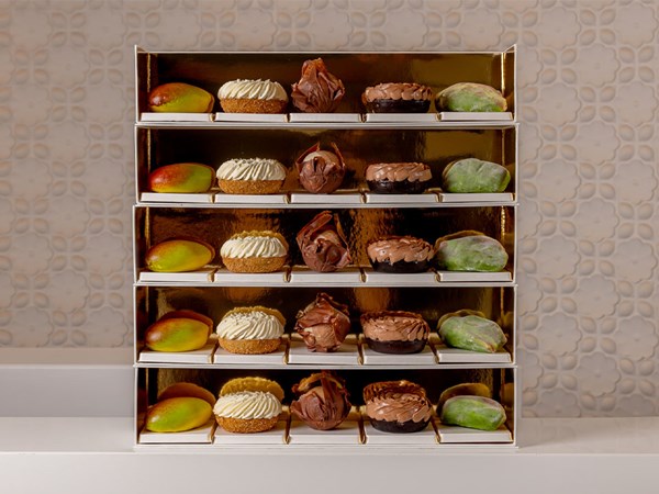 A luxurious tiered presentation of Cedric Grolet’s pastry collection, featuring mango-shaped designs, chocolate swirls, and cream-topped delights, set against a golden backdrop.