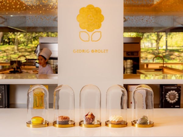 A sleek dessert counter displaying five of Cedric Grolet’s signature creations under individual glass domes, with a branded backdrop and a chef in the background.