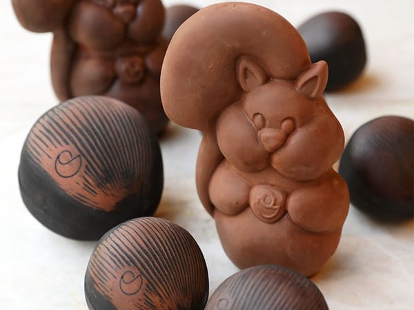 A charming chocolate squirrel sculpture with a bushy tail, surrounded by glossy, nut-shaped chocolate pieces decorated with fine brown and black lines.