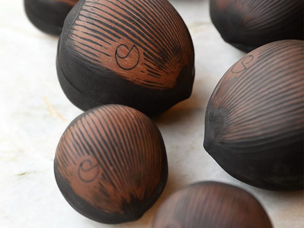 Close-up of dark chocolate nut-shaped confections with fine etched patterns and a subtle monogram detail on each piece, arranged on a marble surface.