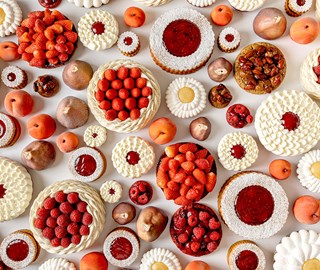 A vibrant flat lay of Cedric Grolet summer pastries, featuring cream-filled tartlets, glazed fruit cakes, and lifelike peach and fig desserts.