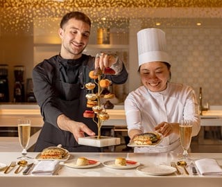 Two pastry chefs present an elegant vertical stand of mini desserts alongside champagne and gourmet snacks at Cedric Grolet’s tasting counter.