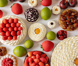 A vibrant overhead composition of seasonal pastries, piped tarts, and lifelike fruit-shaped desserts arranged among fresh limes, cherries, and berries on white.